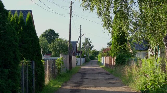 Static wide shot shows narrow dirt lane bordered by wooden utility poles and lush fences between private houses in Daugavpils, Latgale region, Latvia.