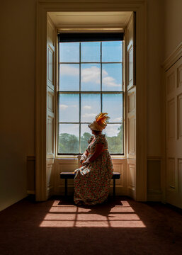 A woman dressed in a vintage period costume with a feathered hat sits on a bench in front of a large window, looking out while attending a historical event.