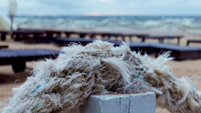 Detail closeup shot focuses on thick frayed nautical rope mounted on white timber post along seaside lounge terrace overlooking stormy Baltic waves in Jurmala.