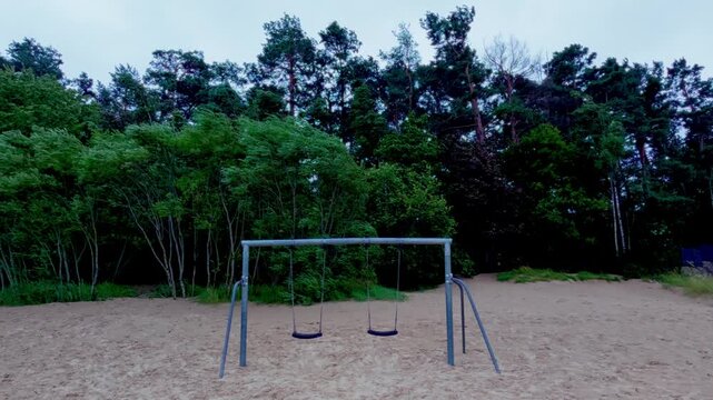 Wide static view captures lonely playground swing setup on Baltic seashore sand dunes with dense pine tree forest blowing in strong coastal gust under overcast evening sky.
