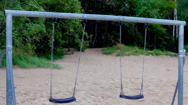 Static wide shot of vacant outdoor playground swing set situated on beach sand with lush green coastal pine woods swaying in strong wind in Jurmala resort.