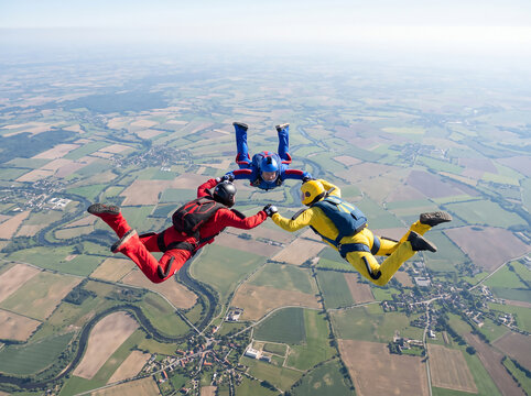 Three skydivers in colorful jumpsuits holding hands in formation freefall over agricultural fields with extreme sport air sports skydiving
