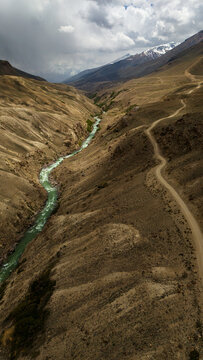 Aerial view of Panj River flowing through the rugged mountain valley with a winding dirt road along the slopes under a cloudy sky in Wakhan, Badakhshan, Afghanistan.
