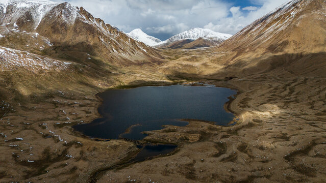 Aerial view of the Pamir Mountains featuring a serene alpine lake surrounded by rugged, snow-capped peaks and vast, textured terrain in Jirgatol, Gorno-Badakhshan Auto, Tajikistan.