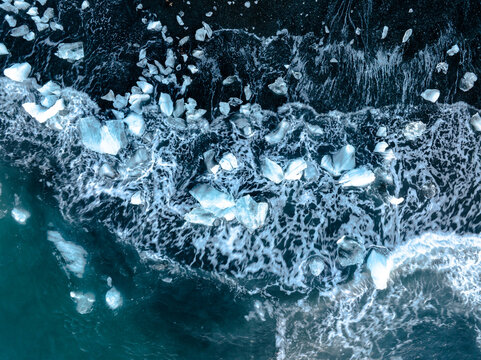 Aerial view of ice chunks scattered on the black sand beach with waves washing over them in Iceland.