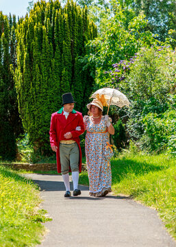 A couple dressed in 19th-century Regency-era clothing walk arm-in-arm along a lush garden path, likely attending a historical reenactment or themed event.