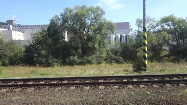 Side view from a moving train riding past an industrial plant and railway tracks on a sunny day