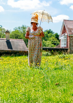 A woman dressed in elegant historical period clothing holds a parasol while walking through a sunny field of yellow wildflowers, likely participating in a themed photoshoot or historical reenactment.
