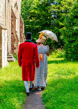 A man in a red frock coat and top hat and a woman in a floral regency dress walk together past a historic stone building, presumably arriving for a period-themed event or wedding.