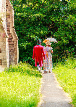 A couple dressed in 19th-century Regency attire, including a top hat, tailcoat, and parasol, walk together along a sunny garden path, likely participating in a historical reenactment event.