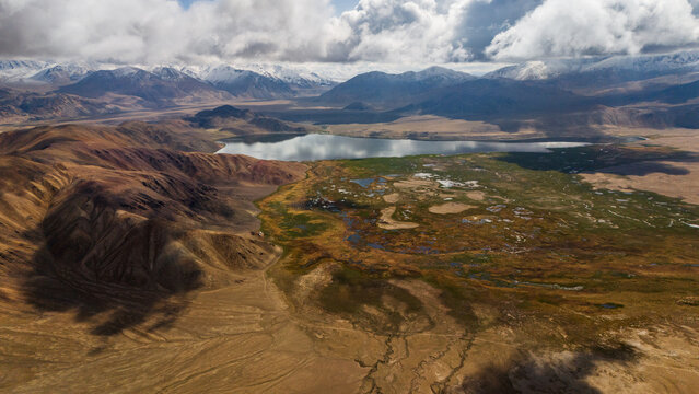 Aerial view of Bulunkul lake surrounded by arid mountain ranges with snow-capped peaks and marshy wetlands under a cloudy sky in Chah Ab, Gorno-Badakhshan Auto, Tajikistan.