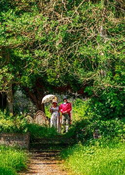 A couple in 19th-century historical attire, featuring a man in a red frock coat and a woman with a parasol, walk through a lush, green cemetery during a historical re-enactment event.
