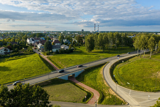 Aerial view of a road junction with a bridge over a path, surrounded by green fields, trees, and residential houses under a blue sky in Kaunas, Kaunas County, Lithuania.