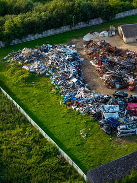 Aerial view of a large scrapyard filled with piles of discarded metal and crushed vehicles, surrounded by green grass and a fence in Kaunas, Kaunas County, Lithuania.
