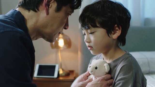 Leaning father holding teddy bear comforting son on bed, in dark sweater, tablet, lamp glowing