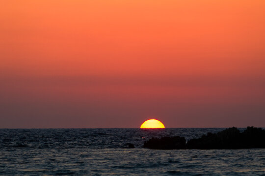 A stunning golden sun setting over the sea at Voidokilia beach, Messenia, Greece, with silhouette rocks and a vibrant orange sky.