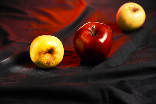 Naturaleza muerta con manzana roja brillante y dos manzanas amarillas sobre tela oscura.
