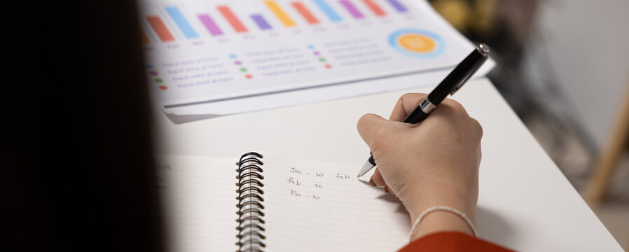 Hand writing business notes beside colorful report chart during office work with focused attention and planning