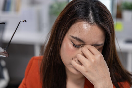 Eye strain woman office fatigue headache glasses stress dark hair red shirt adult shows discomfort while pinching her nose in workplace suggesting tiredness and visual fatigue during computer work