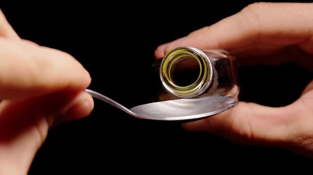 Hands Pour Thick Brown Liquid From a Glass Bottle Into a Metal Spoon Against a Black Background, Showing Careful Measurement and Preparation