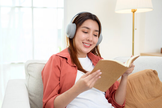 Happy Asian Woman Reading a Book While Wearing Wireless Headphones and Relaxing at Home