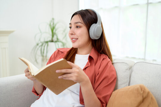 Happy Asian Woman Reading a Book While Wearing Wireless Headphones and Relaxing at Home