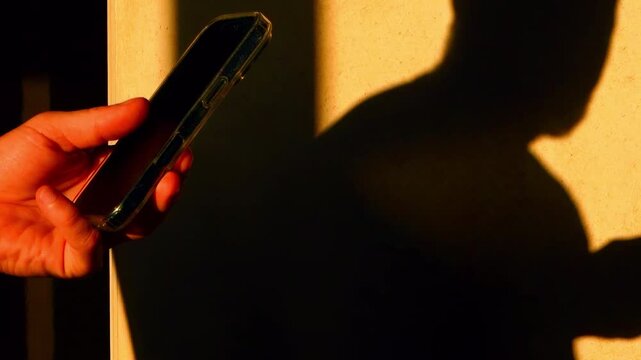 Close-up hands using a smart phone. Casting silhouette shadow on the yellow wall. Doomscrolling
