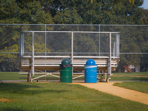 Baseball dugout with aluminum bleachers and green and blue trash cans at a public park sports field