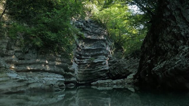 Handheld timelapse of sun rays moving across textured white rocks in Martvili Canyon, Georgia. Calm river water reflects the rugged gorge and lush green foliage.