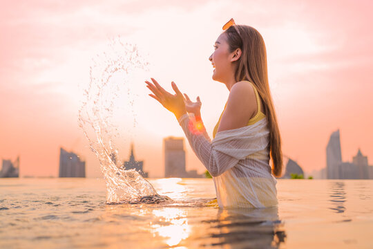 Carefree young woman enjoying summer vacation freedom, splashing water at rooftop infinity pool sunset, expressing pure joy, happiness, vitality, and luxury lifestyle retreat.