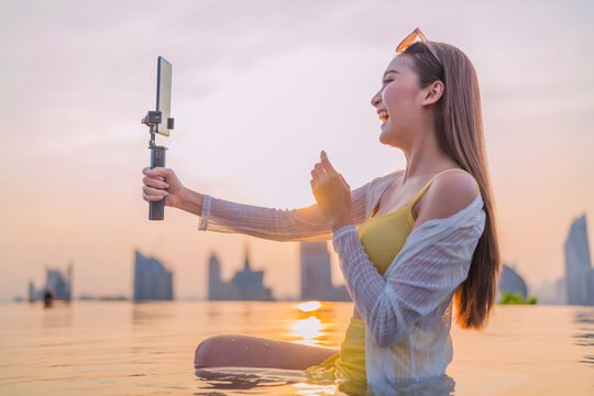 Cheerful young Asian woman vlogging at sunset by a rooftop infinity pool, laughing while recording content on her smartphone gimbal against a glowing city skyline, living the carefree travel lifestyle