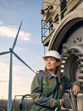 Vertical portrait of a female wind energy technician in a hard hat looking away. Professional woman engineer standing on a platform near a wind turbine
