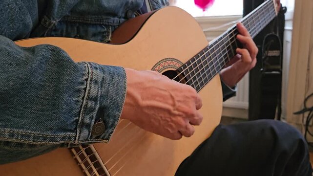 A close-up of a musician s hands playing an acoustic classical guitar at home, focusing on finger placement, the wooden floor, denim jacket, and the intimate atmosphere of practice and concentration.