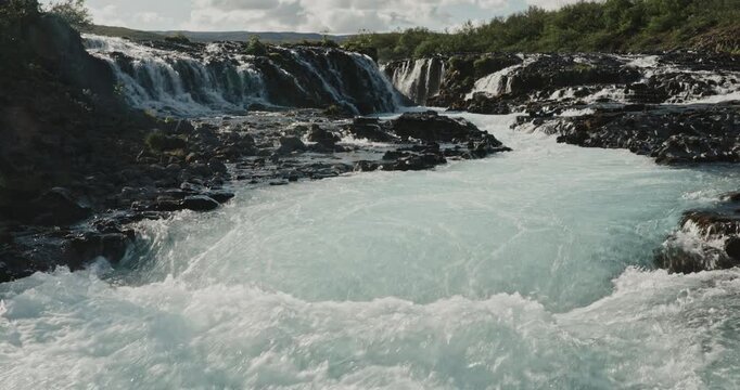Br&uacute;arfoss waterfall cascading through dark volcanic rocks in Iceland, with vivid turquoise glacial water flowing through a rugged natural landscape surrounded by lush green vegetation.