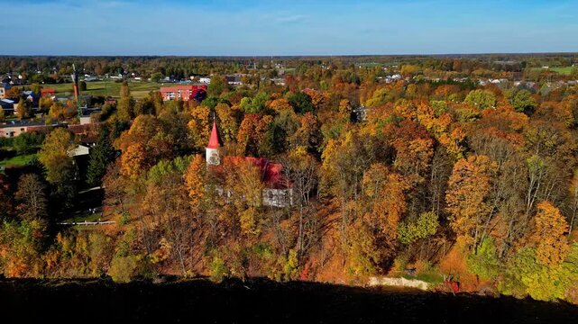 Aerial view of a church with a red roof nestled among vibrant autumn foliage in Kegums, Ogres novads, Latvia.