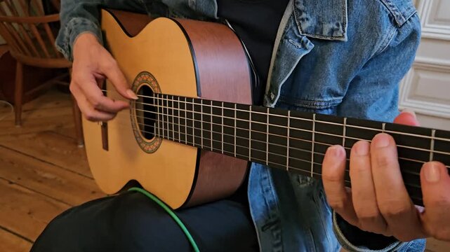 A close-up of a musician s hands playing an acoustic classical guitar at home, focusing on finger placement, the wooden floor, denim jacket, and the intimate atmosphere of practice and concentration.