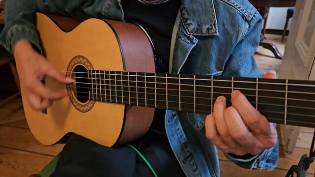 A close-up of a musician s hands playing an acoustic classical guitar at home, focusing on finger placement, the wooden floor, denim jacket, and the intimate atmosphere of practice and concentration.