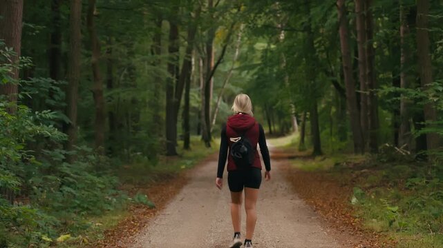 Back view of a Caucasian woman walking along a forest path in summer, surrounded by beautiful dense green trees and lush foliage.