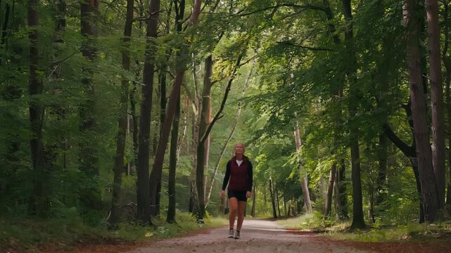 Front view of a caucasian woman walking along a peaceful forest path in summer, surrounded by green trees and soft sunlight filtering beautifully through the foliage.