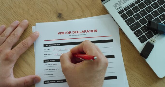 Close-up of a person completing a visitor declaration form at an office desk with a laptop, representing workplace security, registration and access control.