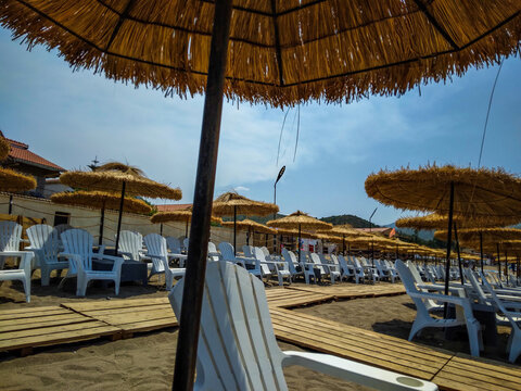 Traditional straw beach umbrellas provide cooling shade over rows of white plastic chairs positioned on wooden boardwalks during a sunny summer afternoon at seaside resort