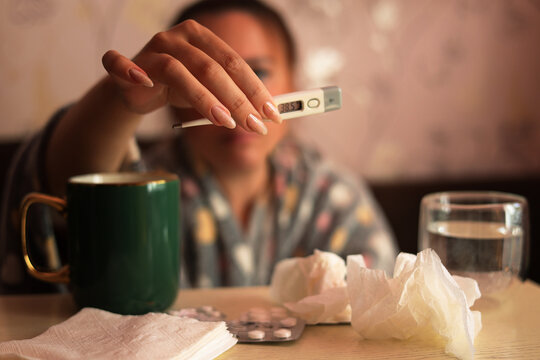 Sick woman showing digital thermometer with fever sitting at table with medicine