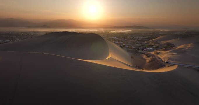 Person trudge up crest of dune in golden light of sunrise at Huacachina, aerial