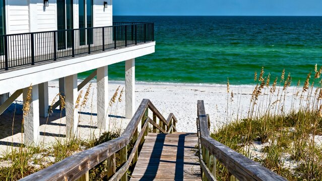A modern elevated beach house in Alabama's Gulf Shores with white stucco and black metal railings, harsh sun casting hard shadows, sugar-white sand and emerald-green water.