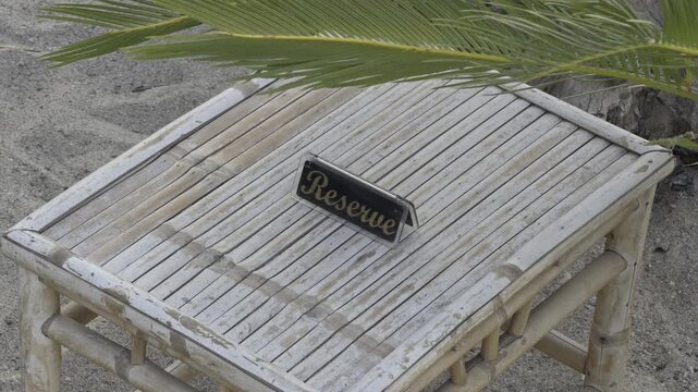 A small reserved sign stands on a rustic wooden bamboo table on the sand shaded by a palm leaf at a beach resort in Chalkidiki Greece
