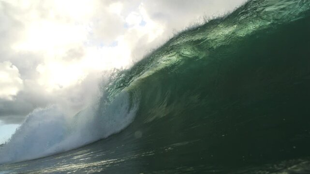 Dark green ocean wave curling beneath bright clouds near water level in smooth slow motion