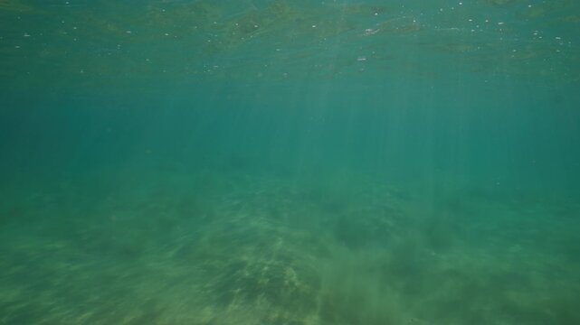 Rippled sand covers a shallow ocean floor beneath clear turquoise water and soft natural light, rays of light gradually descend in columns
