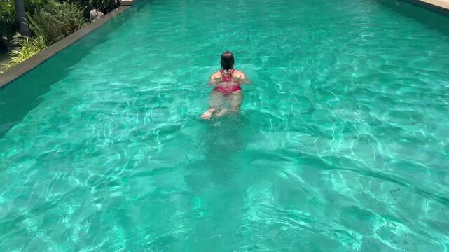 Woman swims in clear pool, making waves as they move. The sun shines brightly on water, reflecting light and creating a playful look. It is a warm day.