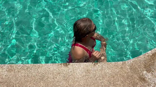 Woman swims in clear pool, making waves as they move. The sun shines brightly on water, reflecting light and creating a playful look. It is a warm day.