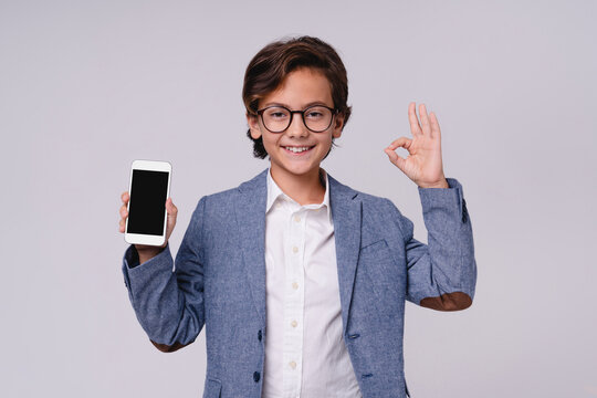 Confident young boy showing okay gesture and holding smartphone isolated on grey background, childhood, technology, communication, digital lifestyle and future businessman concept